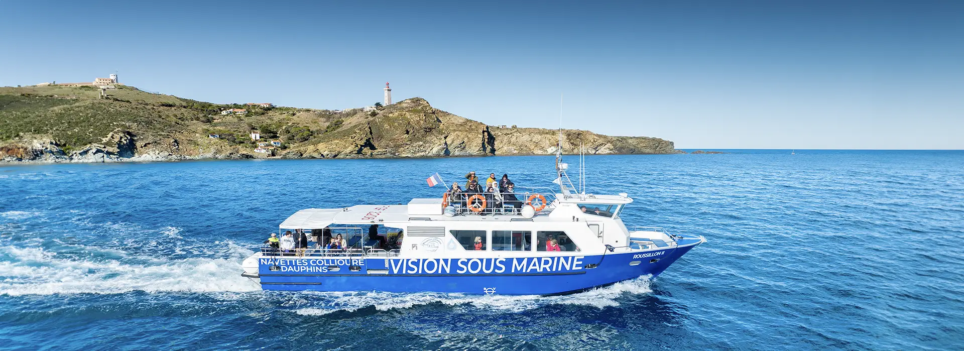 The boat "Roussillon 2" sailing near Cap Béar, with its iconic lighthouse in the background and crystal-clear blue waters.